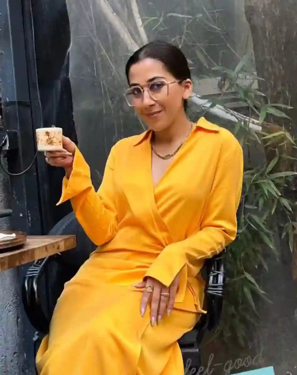 Woman wearing 18k Gold Plated necklace and rings, seated in yellow dress holding a cup at cafe